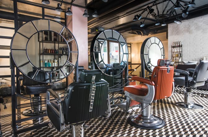 loft style barbershop with coffee area, lounge area, brick walls, stairs, & grey, orange, black chairs. Round tiled mirrors. Black & white tiled carpet. 